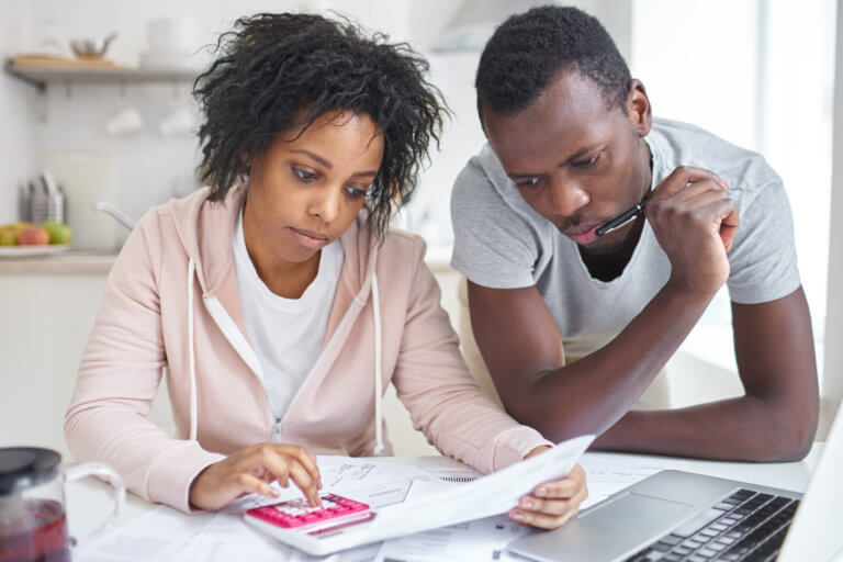 Stressed African-American couple working through paperwork together, calculating expenses, trying to save some money, managing family finances, sitting at kitchen table with laptop and calculator